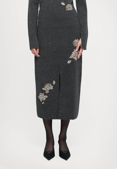 Vestido largo de punto gris oscuro con adornos florales plateados, abertura delantera, combinado con medias transparentes negras y tacones negros de punta.