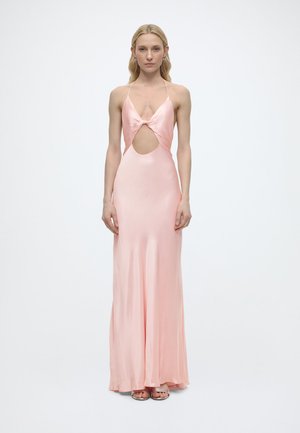 Kvinde iført en gulvlængde, silkeblød lyserød halterkjole med en udskæring foran, stående mod en ensfarvet hvid baggrund.
