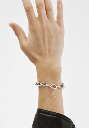 Mano con pulsera de cadena plateada con un colgante en forma de "L", sobre un fondo blanco liso, con una manga de cuero negro.