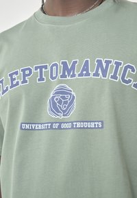 Green cotton t-shirt featuring bold navy text "LEPTOMANICS" and "UNIVERSITY OF GOOD THOUGHTS," with an illustration of two hands forming a circle.
