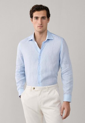 Joven con camisa azul clara abotonada y pantalones blancos, de pie contra un fondo liso, manos en los bolsillos, expresión neutral.