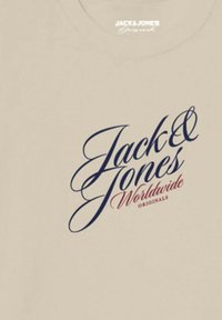 Beige cotton t-shirt with a large, blue cursive "Jack & Jones" logo and red "Worldwide Originals" text beneath it.