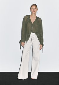 Blusa verde oliva trasparente con scollatura a V e bottoni davanti, abbinata a pantaloni bianchi a gamba larga con dettaglio allacciato e vita alta.