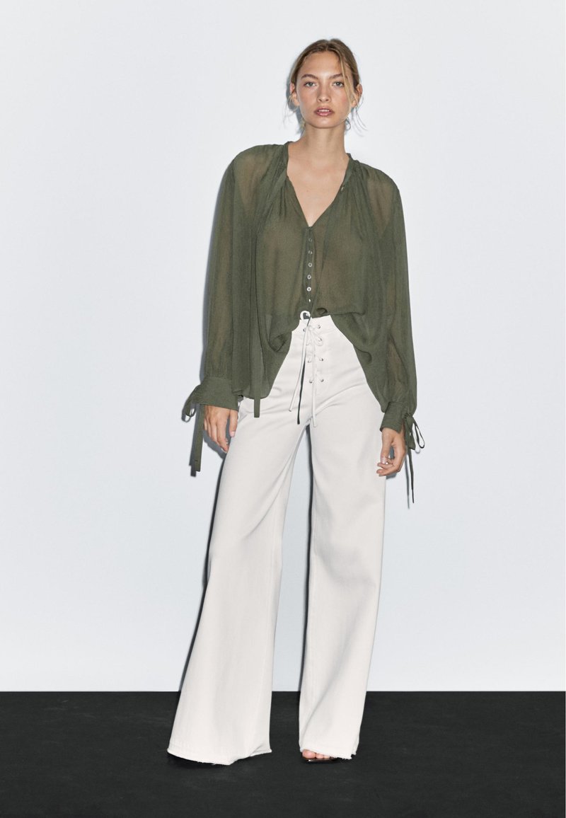 Blusa verde oliva trasparente con scollatura a V e bottoni davanti, abbinata a pantaloni bianchi a gamba larga con dettaglio allacciato e vita alta.