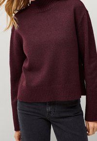 Pull en tricot bordeaux avec un col montant, des poignets et un ourlet côtelés ; coupe décontractée avec une texture douce, associé à un jean foncé.