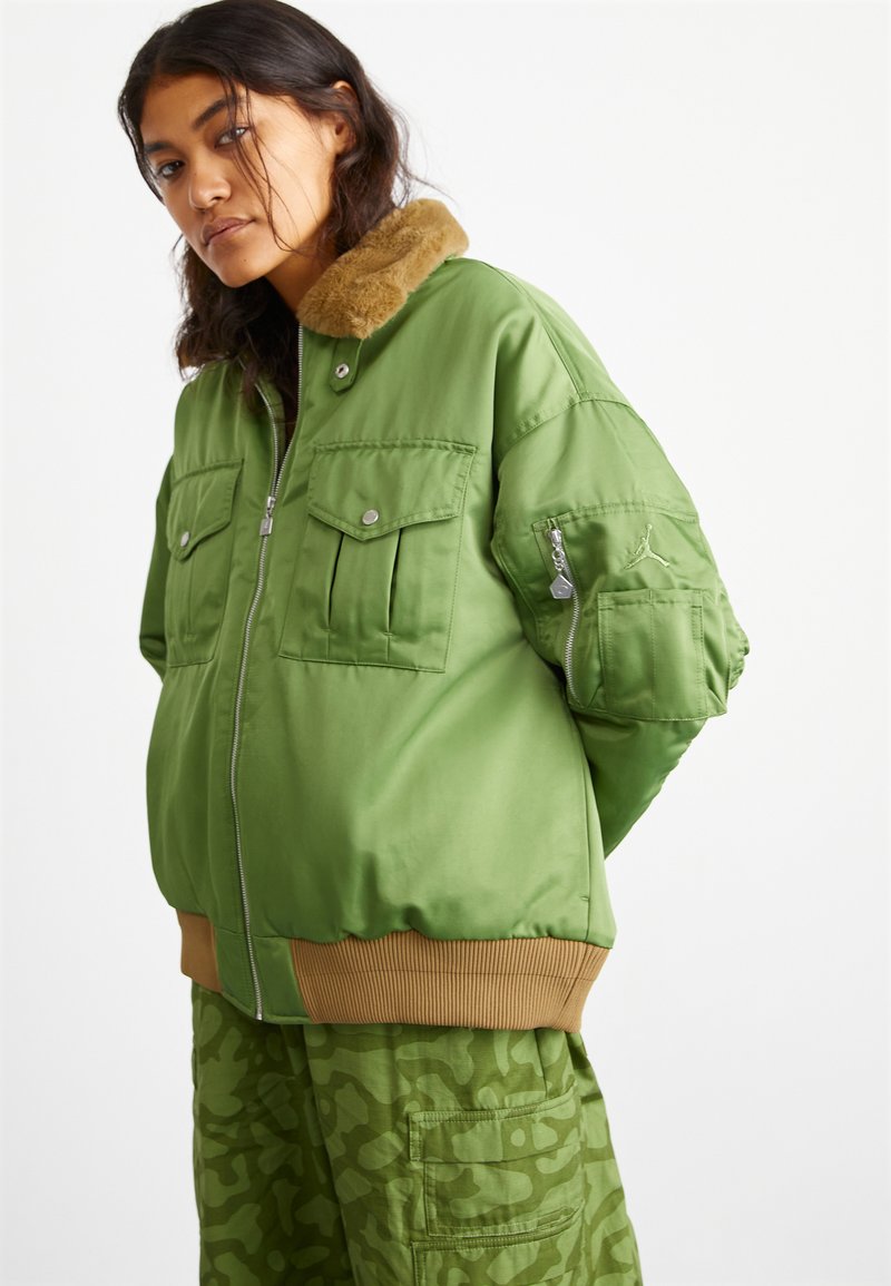 Giacca bomber verde con colletto in morbido pelliccia, chiusura con zip, due tasche frontali e polsini a costine. Indossata con pantaloni cargo mimetici coordinati.