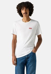 Mand med krøllet hår iført en hvid Levi's t-shirt og blå jeans, stående med den ene hånd i lommen foran en ensfarvet baggrund.
