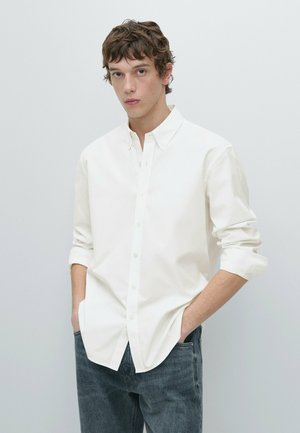 Junger Mann mit kurzen braunen Haaren, der ein weißes Hemd mit Knöpfen und blaue Jeans trägt, steht mit den Händen in den Taschen vor einem einfarbigen Hintergrund.