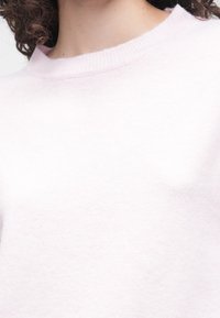 Svetlo roza pleten pulover z okroglim izrezom in mehkim, teksturiranim zaključkom. Tkanina je videti gladka in lahka.