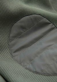 Gros plan sur un tissu vert olive texturé avec un patch ovale en cuir lisse cousu dessus.