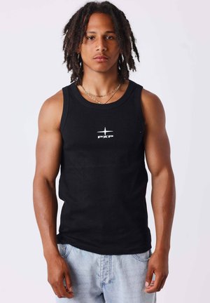Giovane uomo con dreadlocks di media lunghezza, indossa una canottiera nera con logo bianco e jeans blu chiaro, in piedi davanti a uno sfondo neutro.