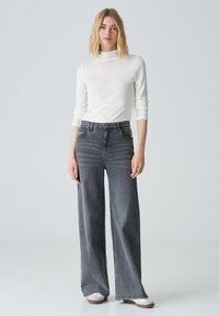 Weite graue Jeans aus Denim, mit hoher Taille, Vordertaschen und subtilen Ausbleichungen. Kombiniert mit einem eng anliegenden weißen Langarmshirt.