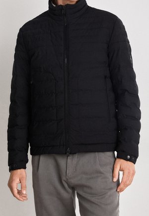 Hombre que lleva una chaqueta negra acolchada con cremallera frontal y puños abotonados, combinada con pantalones grises, sobre un fondo liso.