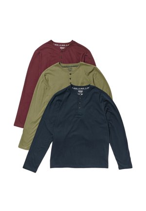 Mand stående iført en mørkeblå langærmet henleytrøje, beige kordbuksere og blå sneakers mod en ensfarvet lys baggrund.
