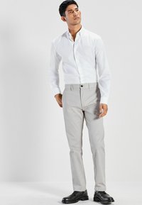 Weißes Hemd mit langen Ärmeln, kombiniert mit hellgrauen Hosen und schwarzen Schuhen. Das Outfit zeichnet sich durch eine körperbetonte Passform und eine glatte Textur aus.