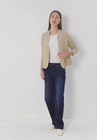 Frau steht vor einer weißen Wand, trägt einen beigen Blazer, ein weißes Hemd, dunkelblaue weite Jeans und weiße Sneaker.
