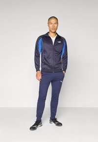 Puma treningsdrakt i marineblått med blå detaljer. Jakke med glidelås, tettsittende bukser og svarte joggesko. Glatt tekstur med logo-detalj.