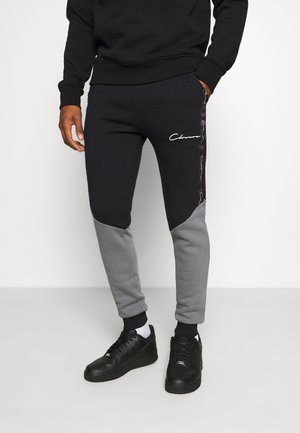 Mann trägt schwarze und graue Jogginghose mit "Classic"-Logo am oberen Oberschenkel, schwarze Turnschuhe, weiße Socken und schwarzen Pullover.