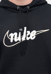 Černá mikina s kapucí s velkým bílým vyšívaným logem "nike". Šňůrky s koncovkami, měkká textura a volný střih.
