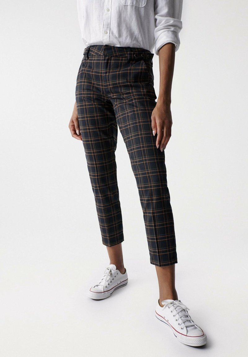 Personne portant un pantalon slim noir à carreaux avec des lignes orange, une chemise blanche à manches longues retroussées et des baskets blanches décontractées, debout sur un fond blanc.