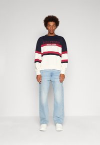 Felpa a strisce blu navy, bianca e rossa con logo "HILFIGER NEW YORK". Abbinata a jeans blu chiaro e sneakers bianche.
