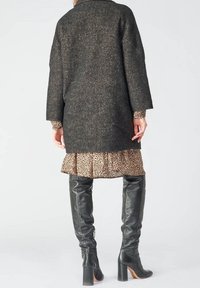Cappotto di lana a zig-zag nero con spalle basse, indossato sopra un vestito beige con stampa leopardata e orlo arricciato, abbinato a stivali in pelle al ginocchio.
