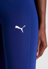 Legginsy Puma w głębokim niebieskim kolorze, z białym logo skaczącego kota. Materiał jest gładki z lekkim rozciągnięciem, widoczne szwy boczne.