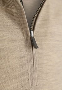 Beige pullover met een ritskraag, gebreide textuur, voorzien van een zilveren rits met 'Suitable' branding en subtiele stikseldetails.