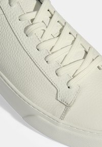 White leather sneaker featuring textured surface, flat white laces, and a rubber sole with a subtle tread pattern. No visible branding.