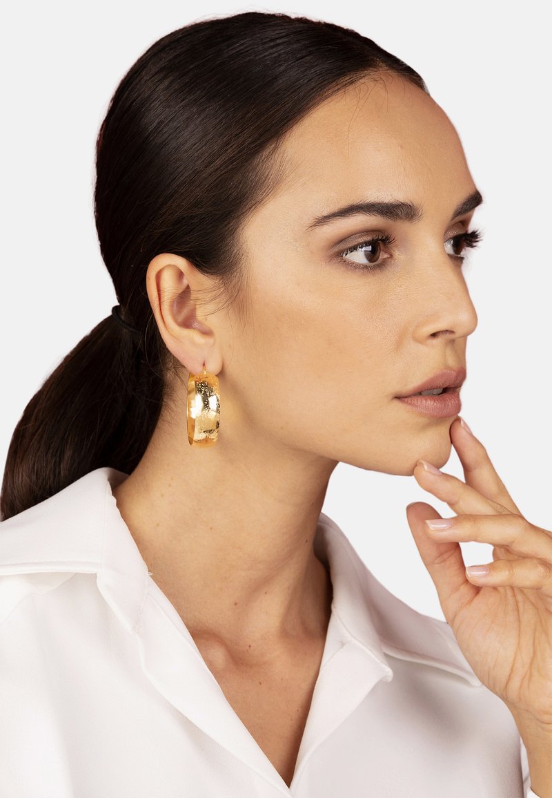 Pendientes de aro de oro con una superficie texturizada, mostrando un acabado pulido. La modelo lleva una camisa blanca con cuello y posa de forma pensativa.