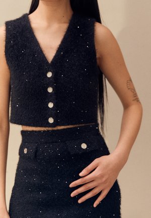 Femme portant un crop top noir texturé sans manches et une jupe taille haute assortie avec des boutons argentés et de subtils éclats.