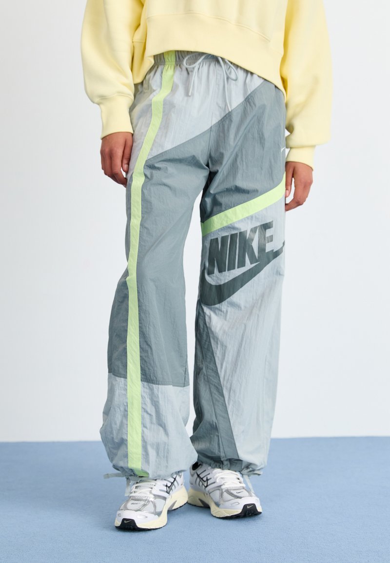 Lightweight, grey track pants with green accents, featuring a large Nike logo and a drawstring waist. Textured fabric with layered designs.