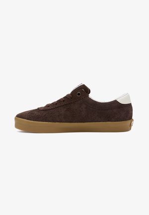 Niski brązowy zamszowy sneaker z gumową podeszwą, brązowymi sznurowadłami, metalowym oczkiem i białą nakładką na pięcie przedstawiony z profilu bocznego.