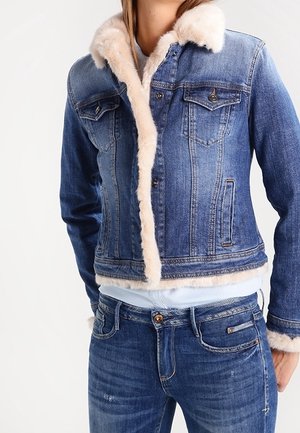 Frau trägt blaue Jeansjacke mit beigeem Pelzfutter und passende blaue Jeans vor weißem Hintergrund.