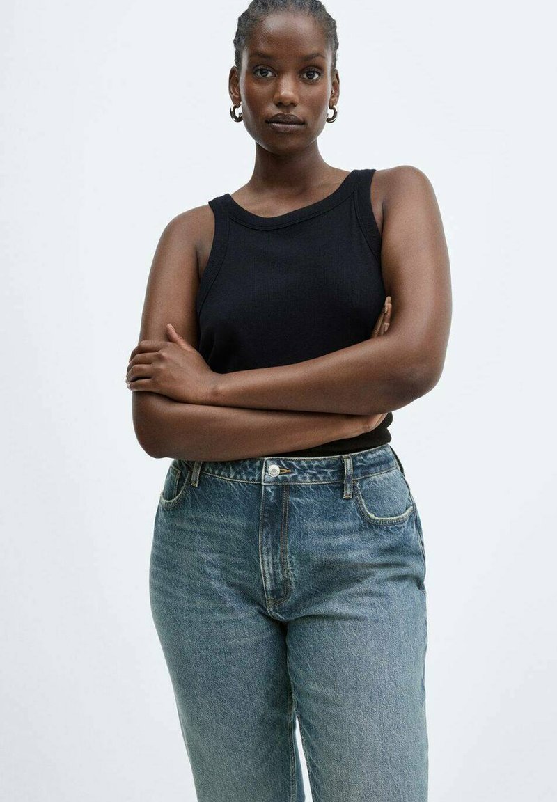 Schwarzes ärmelloses Tanktop, kombiniert mit hochgeschnittenen blauen Jeans, die einen klassischen Schnitt und eine abgenutzte Textur aufweisen. Das Model steht mit gekreuzten Armen.