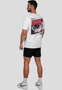 Camiseta blanca de algodón con un gráfico en la parte posterior con un tema de casino, que incluye dados y un teclado, con acentos rojos y negros. Llevada con pantalones cortos negros.