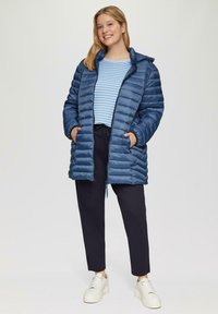 Marineblauwe gewatteerde jas met een capuchon, rits aan de voorkant en twee zijzakken; gedragen over een lichtblauwe en witte gestreepte shirt en donkere broek.