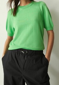 Korte mouwen, lichtgroene gebreide top met een ronde hals en een ontspannen silhouet. Gecombineerd met zwarte, elastische broeken met zakken.