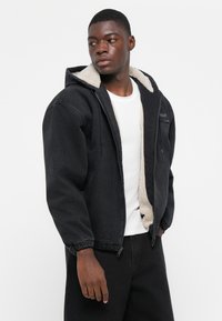 Svart zip-up jakke med hette kledd i lammeskinn. Har elastiske mansjetter og en lomme foran; glatt ytre overflate med myk innvendig tekstur.