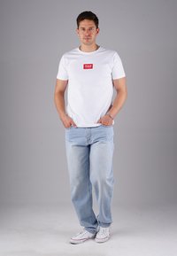 Mann steht mit den Händen in den Taschen, trägt ein weißes T-Shirt mit rotem Logo, hellblaue Jeans und weiße Sneakers vor einem grauen Hintergrund.