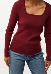 Burgunderfarbener gerippter Strickpullover mit quadratischem Ausschnitt, langen Ärmeln und taillierter Passform, kombiniert mit blauen Jeans. Auffällige Textur- und Farbkontraste.