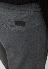 Grå tygbyxor med synlig textur, med en svart läderlogotags och sidofickor med dragkedja. Elastisk midja för komfort.