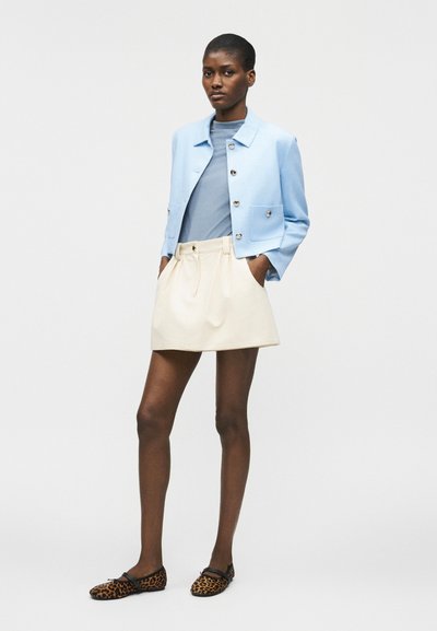 Γυναίκα με ανοιχτό μπλε cropped τζάκετ, μπλε τοπ, κρεμ μίνι φούστα και λεοπάρ φλατ παπούτσια, στέκεται με τα χέρια στις τσέπες.