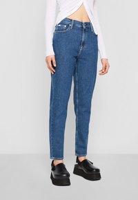 Džínové kalhoty v středním odstínu, vysokým pasem a rovnými nohavicemi. Kombinované s černými platformovými nazouvacími botami. Bílé dlouhé tričko s knoflíky.