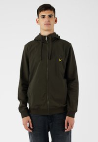Felpa con zip verde oliva, con cappuccio regolabile, polsini e orlo a costine, e un piccolo logo giallo sul petto, indossata sopra jeans scuri.