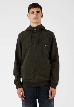 Felpa con zip verde oliva, con cappuccio regolabile, polsini e orlo a costine, e un piccolo logo giallo sul petto, indossata sopra jeans scuri.