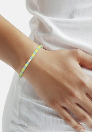 Wrist wearing a beaded bracelet with alternating blue and yellow beads and a small gold square charm, resting on white fabric clothing.