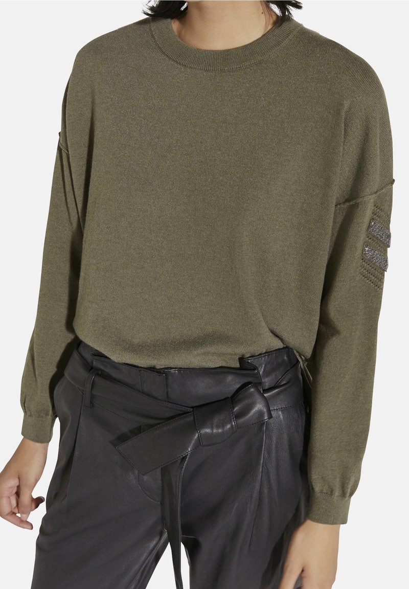 Pull tricoté vert olive avec un col rond et une coupe ample, présentant des rayures métalliques texturées sur les manches. Associé à un pantalon en cuir noir.