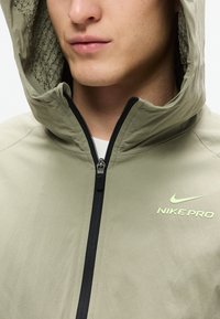 Μπουφάν Nike Pro σε ανοιχτό ελαιδί χρώμα, με μαύρο φερμουάρ, υφασμάτινη επένδυση κουκούλας με υφή και ανακλαστική λεπτομέρεια λογότυπου στο στήθος.