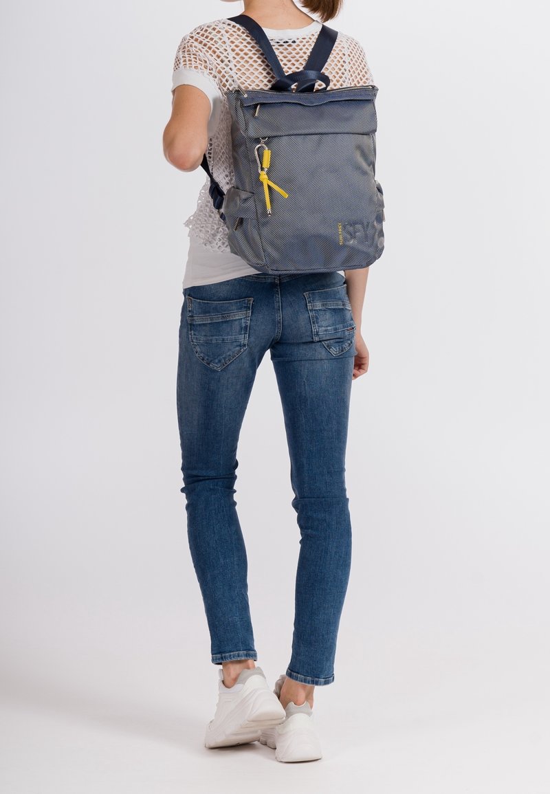 Personne portant un jean bleu, des baskets blanches et un haut blanc en mesh à manches courtes, portant un sac à dos gris avec une fermeture éclair jaune.